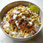 A close-up of a finished Street Corn Chicken Rice Bowl with sliced grilled chicken, corn, white rice, cotija cheese, and lime wedges.