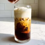 A tall glass of Vanilla Sweet Cream Cold Foam Cold Brew showing distinct layers of dark coffee, ice, and thick white foam.