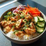 A close-up of a Bang Bang Chicken Bowl featuring tender chicken pieces coated in creamy sauce, served over rice with fresh broccoli, shredded carrots, cucumber slices, and red onion.