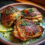 Three crispy baked chicken thighs glistening with herb butter and lemon slices in a bowl.