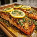 Close-up of perfectly baked salmon fillets with lemon slices and fresh herbs on a baking sheet.