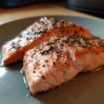 Two perfectly baked salmon fillets seasoned with herbs and spices on a grey plate, showcasing the moist and flaky texture of the salmon.