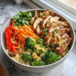 A healthy bowl filled with quinoa, grilled chicken, broccoli, shredded carrots, red bell pepper, and cilantro, drizzled with sauce.