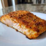 A close-up of a perfectly cooked Parmesan crusted salmon fillet on a white plate, showing its flaky texture.