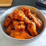 A bowl filled with glossy Buffalo Style Wings coated in a vibrant orange Tangy Butter Sauce and sprinkled with herbs.