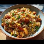 A close-up bowl of Chicken Fried Rice with Extra Crispy Bits, featuring rice, chicken pieces, scrambled egg, peas, and green onions.
