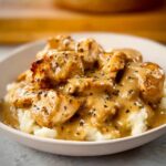 A bowl of creamy mashed potatoes topped with tender pieces of Comfort Style Chicken with Creamy Gravy, drizzled with rich gravy and herbs.