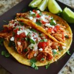 Two delicious tacos filled with shredded Fast Salsa Chicken, topped with cheese, salsa, and cilantro, served with lime wedges.