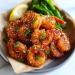 A plate of glistening Honey Sriracha Shrimp coated in a sticky glaze, sprinkled with sesame seeds and green onions, served with broccoli and a lemon wedge.