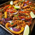 A close-up of colorful Sheet Pan Chicken Fajitas with Peppers, onions, and lime wedges on a baking sheet.
