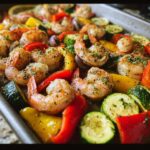 A close-up of a sheet pan filled with cooked shrimp, zucchini, bell peppers, and red onion, seasoned with herbs.
