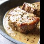 Close-up of two thick skillet pork chops smothered in a creamy Dijon sauce, garnished with herbs and pepper.