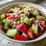 A close-up of a vibrant Greek Salad Recipe bowl filled with chopped cucumbers, tomatoes, red onion, feta cheese, and olives.