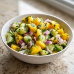 A close-up of a vibrant Mango Cucumber Salad Recipe in a white bowl, featuring diced mango, cucumber, red onion, and cilantro.